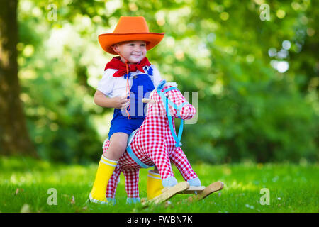 Little boy dressed up as cowboy playing with his toy rocking horse in a summer park. Kids play outdoors. Children in Halloween Stock Photo