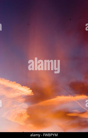 A jet airliner is dwarved by huge clouds as it flies through a dramatic sunset colouring the sky deep red, orange, purple and pink and leaving the jet only visible as a tiny silhouette. Stock Photo