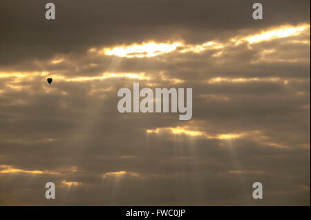 The tiny silhouette of a hot air ballon dwarfed byt he surrounding sky, clouds and rays of sunlight. Stock Photo