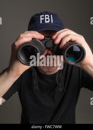 Photograph of an FBI agent in black T-shirt and baseball cap looking through binoculars. Stock Photo