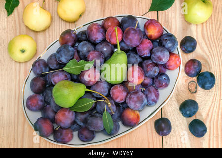 On the surface of the wooden table is a ceramic dish filled with large Mature plums, pears. Next to them are apples. Stock Photo