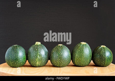 Five raw eight ball squashes in a row on a natural wooden board against a black textured wooden background. Stock Photo