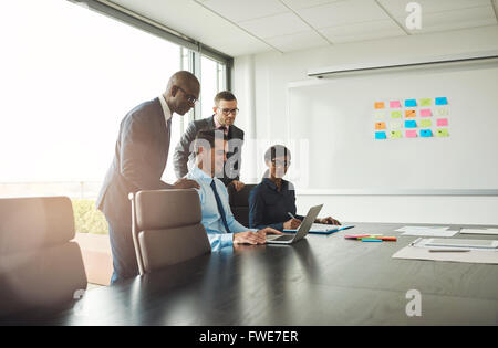 Group of four Black and white young business people at conference table looking at laptop with sticky note chart on board Stock Photo