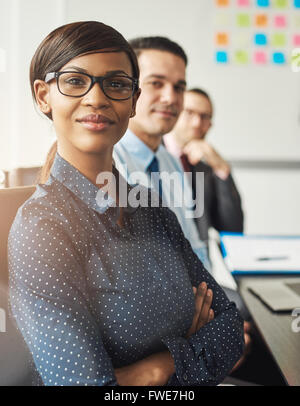 Confident smiling business woman wearing eyeglasses and white polka dotted shirt seated with folded arms beside male co-workers Stock Photo