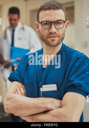 Serious confident young surgeon wearing glasses and surgical scrubs standing in a hospital with folded arms looking intently at Stock Photo