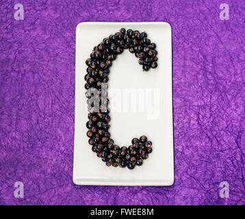 Letter C made of rows of blackcurrants on a rectangular white plate against a purple background. Stock Photo