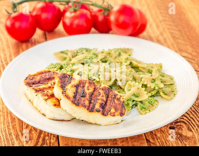 Lateral view of grilled halloumi cheese with pesto farfalle with a vine of tomatoes on a wooden table. Stock Photo
