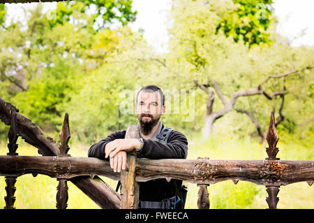 Bearded young man leaning on an old carved wooden fence. Stock Photo