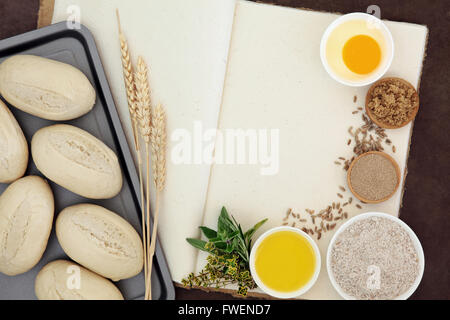 Fresh dough bread rolls with baking ingredients, wheat sheaths and herbs on old hemp notebook over lokta background. Stock Photo