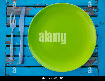 An empty green plate on a weathered blue table along with a fork and knife sits ready to be served a meal for eating. Stock Photo