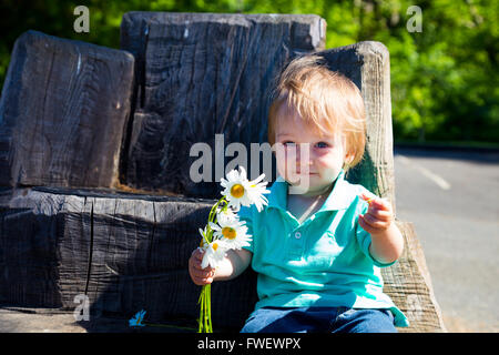 A young boy plays with some flowers while sitting on a hand carved seat made from an old stump. Stock Photo