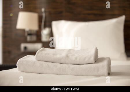 Stacked clean white bath towels on the bed sheets in hotel room, close-up Stock Photo