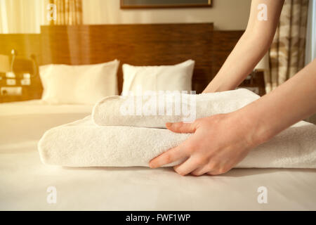 Close-up of hands putting stack of fresh white bath towels on the bed sheet. Room service maid cleaning hotel room. Lens flair Stock Photo