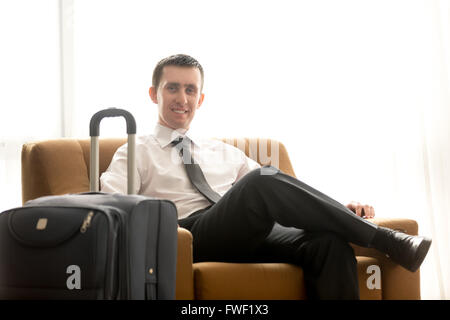 Portrait of handsome smiling young business man with luggage sitting in armchair in hotel room Stock Photo