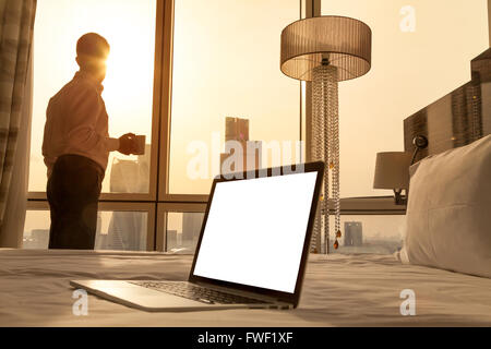 Laptop computer on white bed sheets in cozy room with copy space blank screen. Young businessman with cup of coffee standing Stock Photo
