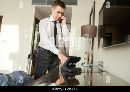 Young traveler businessman wearing white shirt and necktie making call after arriving in the hotel room with his luggage Stock Photo