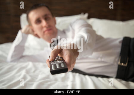 Young businessman wearing white shirt and necktie lying on the bed in the hotel room with remote control in his hand Stock Photo