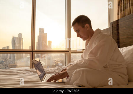Portrait of handsome young businessman wearing white bath robe sitting on the bed with laptop in modern room with sunset Stock Photo