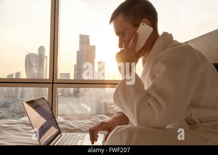 Portrait of handsome young businessman wearing white bath robe sitting on the bed with laptop and cellphone in modern room Stock Photo