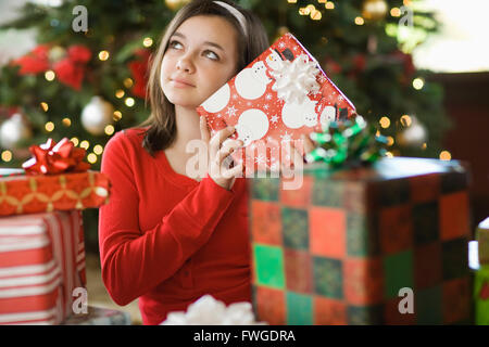 A girl by a Christmas tree shaking a present trying to guess what it is. Stock Photo