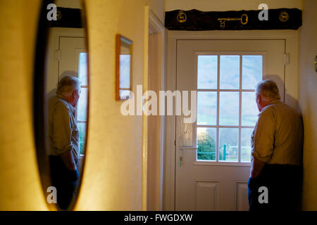 An elderly man standing by a door looking out through the window panes. Stock Photo