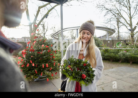 A woman holding a Christmas decorated wreath at a garden centre. Christmas trees in the background. Stock Photo