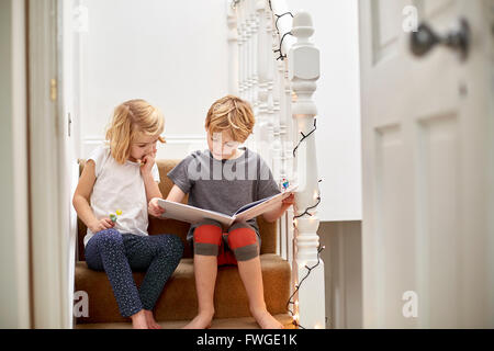 Two children seated side by side on the stairs at home, reading a book together. Stock Photo