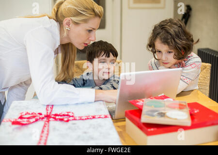 Two children and their mother looking at a laptop computer screen around a kitchen table. Stock Photo