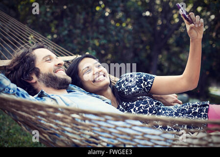 A couple, a young man and woman lying in a large hammock in the garden, taking a selfy of themselves.