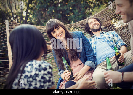 A group of friends lounging in a large hammock in the garden having a beer. Stock Photo
