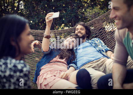 A group of friends lounging in a large hammock in the garden having a beer, and taking a selfie. Stock Photo