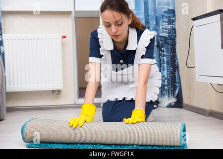Maid or housekeeper doing the cleaning kneeling down and rolling up a small carpet to clean the tiles underneath in a house or hotel. Stock Photo
