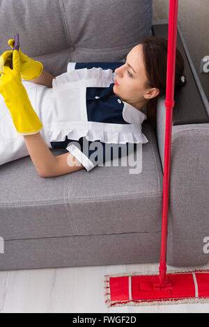 Lazy maid or housekeeper taking a break relaxing on a sofa with her mop beside her reading a text message on her mobile phone, view from above. Stock Photo