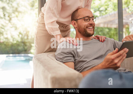 Indoor shot of a smiling man sitting on sofa using digital tablet with woman standing behind him. Stock Photo