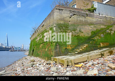 Wapping & River Thames foreshore at low tide showing old base walls of warehouse buildings with Tower Bridge and Shard beyond England UK Stock Photo