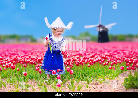 Adorable toddler girl wearing Dutch traditional national costume dress and hat playing in field of blooming tulips next windmill Stock Photo