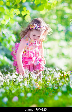 Adorable curly toddler girl in a pink summer dress playing with Easter eggs during egg hunt in a sunny garden Stock Photo