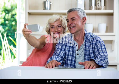 Senior couple taking selfie through mobile phone while sitting in kitchen Stock Photo