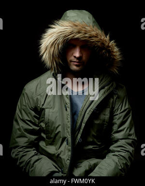 Low key studio portrait of suspicious young adult caucasian model wearing winter coat with hood on. Isolated on black. Stock Photo
