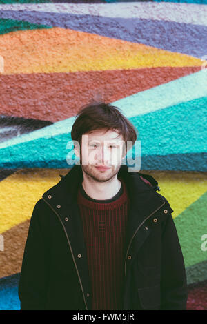 Young caucasian man looking at the camera, wearing warm/casual clothes.  Colour urban wall in the background. Stock Photo