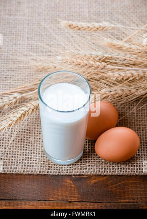 Glass of milk, two eggs and wheat ears lie on the napkin from a burlap on the old wooden table Stock Photo