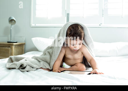 Baby boy covered with towel playing with digital tablet on bed Stock Photo