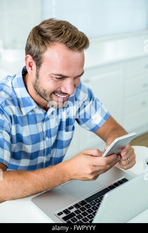 Man smiling while using cellphone by laptop at table in kitchen Stock Photo