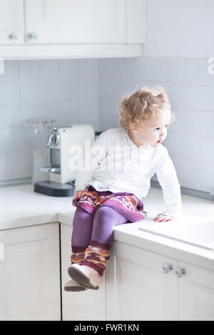 Adorable toddler girl sitting in a white kitchen on the counter top Stock Photo