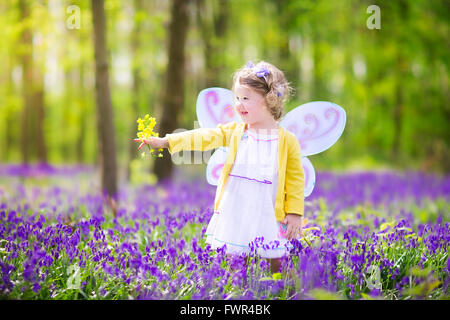 Adorable toddler girl with curly hair wearing a fairy costume with purple wings and yellow dress is playing in beautiful forest Stock Photo