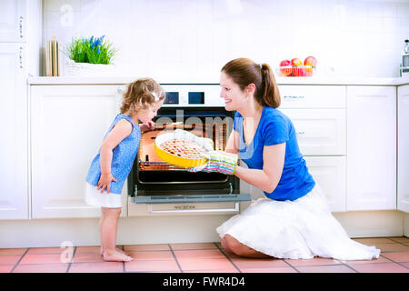 Young happy mother and her adorable curly toddler daughter wearing blue dress baking a pie together in an oven in sunny kitchen Stock Photo