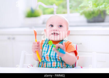 Sweet laughing baby boy sitting in a white sunny kitchen with a garden view window waiting for lunch Stock Photo