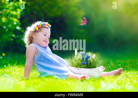 Cute little toddler girl with curly hair wearing a blue summer dress having fun watching a butterfly and flowers Stock Photo