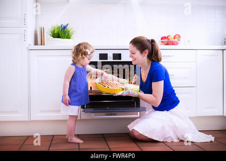 Mother and child bake a pie. Young woman and her daughter cook in a white kitchen. Kids baking pastry. Children helping to make Stock Photo