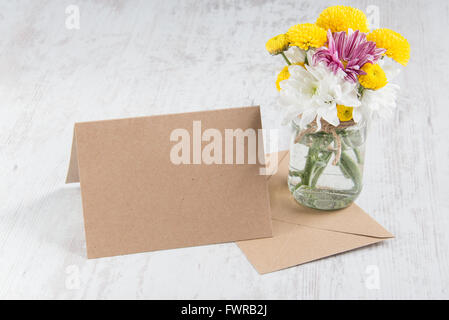 Spring flowers bouquet in a jar vase with kraft card note and envelope on a white wood rustic background Stock Photo